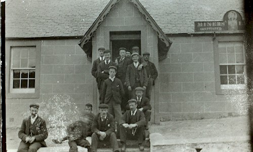 A black and white photo shows 14 men dressed in suits with black boots and flat caps. They sit and stand on the steps of a stone building with a slate roof.