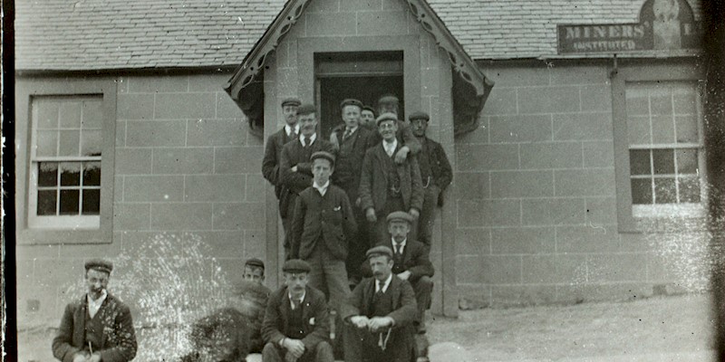 A black and white photo shows 14 men dressed in suits with black boots and flat caps. They sit and stand on the steps of a stone building with a slate roof.