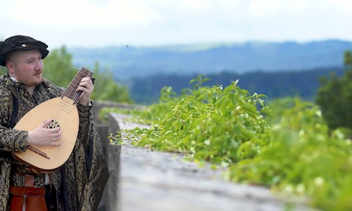 A man in period costume playing a lute while looking over a wall with lovely views of greenery and hills