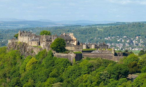 A photo from the air of Stirling Castle. In the foreground is a heavily wooded hill, in the background fields and trees with hills on the horizon. The castle is imposing, with buildings and turrets behind a grey stone wall.