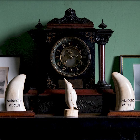 An old clock on a fireplace, along with a whale tooth and a penguin carving