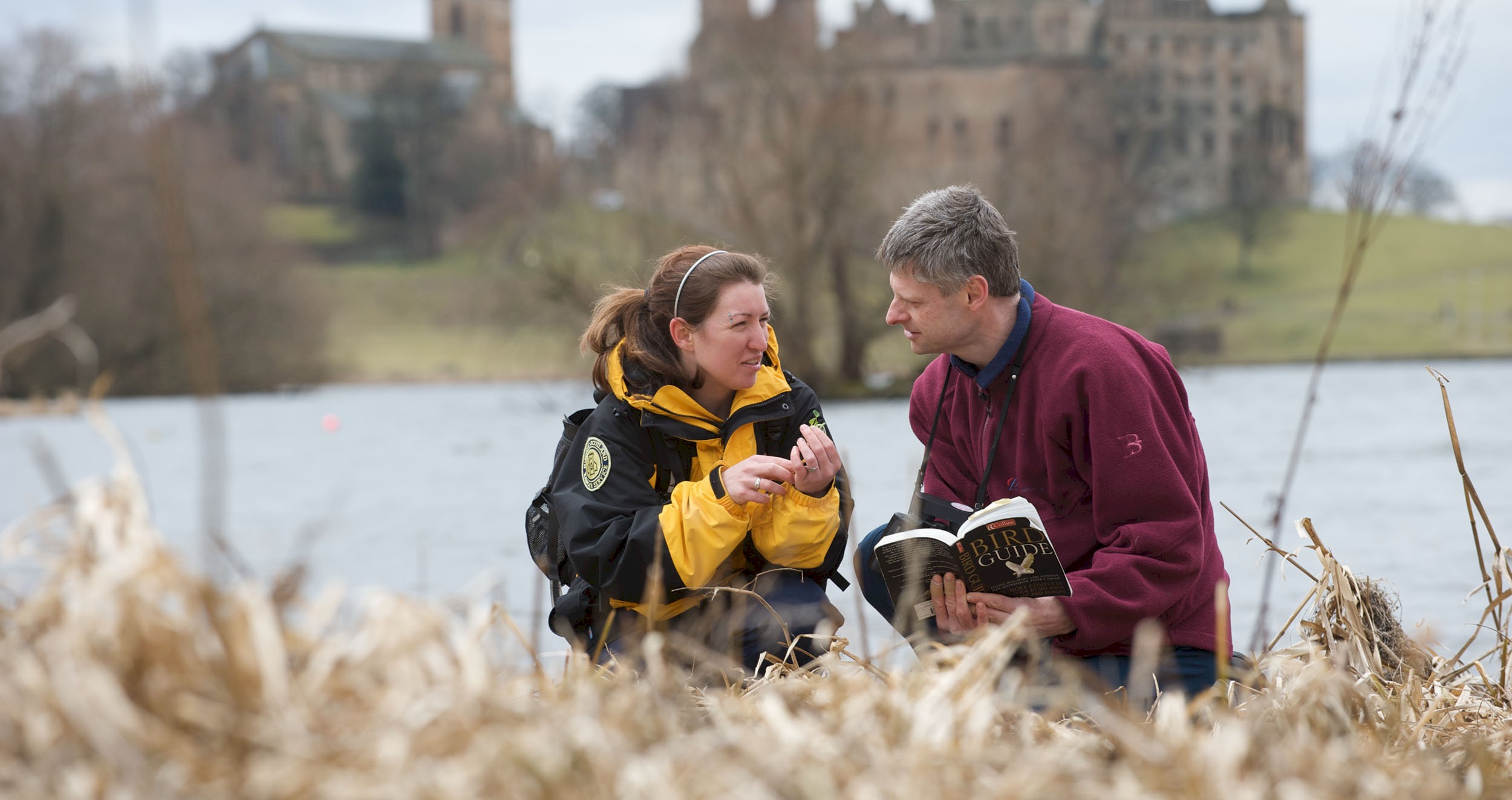 A Ranger with a visitor crouched down by the loch looking at some wildlife and Linlithgow Palace in the background