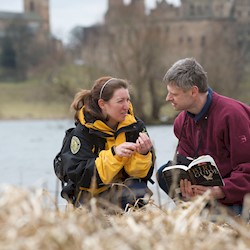 A Ranger with a visitor crouched down by the loch looking at some wildlife and Linlithgow Palace in the background