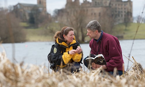 A Ranger with a visitor crouched down by the loch looking at some wildlife and Linlithgow Palace in the background