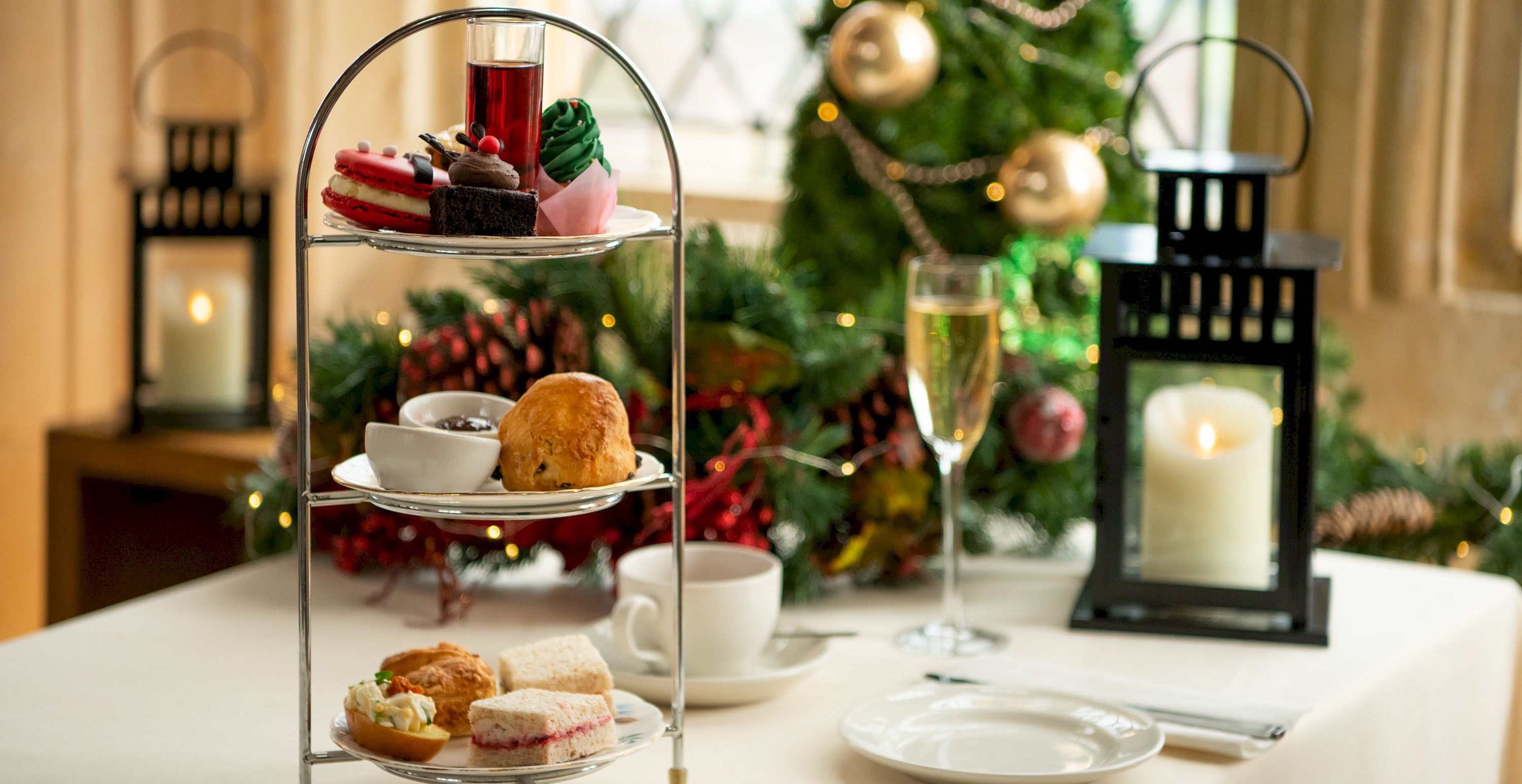 Cake stand with a festive selection for afternoon tea along with a pot of tea, a glass of prosecco and lantern candles 