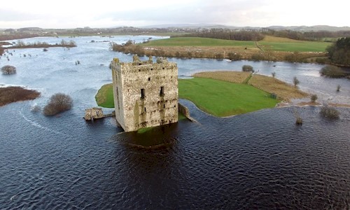 An aerial view of a square-shaped ruined castle on an overcast day. The castle grounds and surrounding fields have been inundated by floodwater, cutting the site off from neighbouring farmland. 