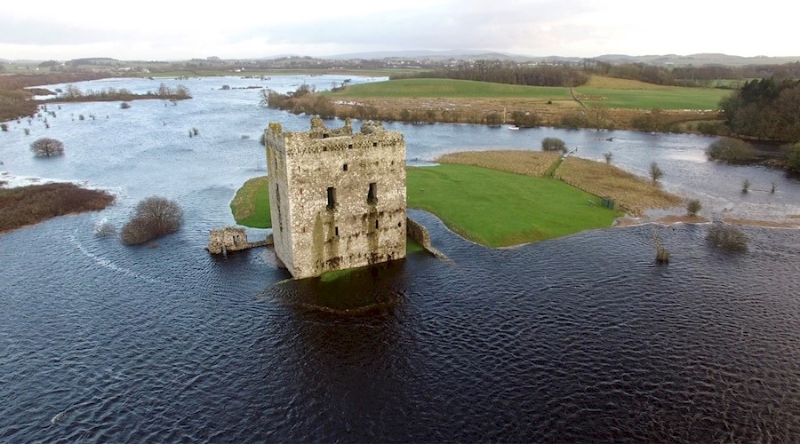 An aerial view of a square-shaped ruined castle on an overcast day. The castle grounds and surrounding fields have been inundated by floodwater, cutting the site off from neighbouring farmland. 