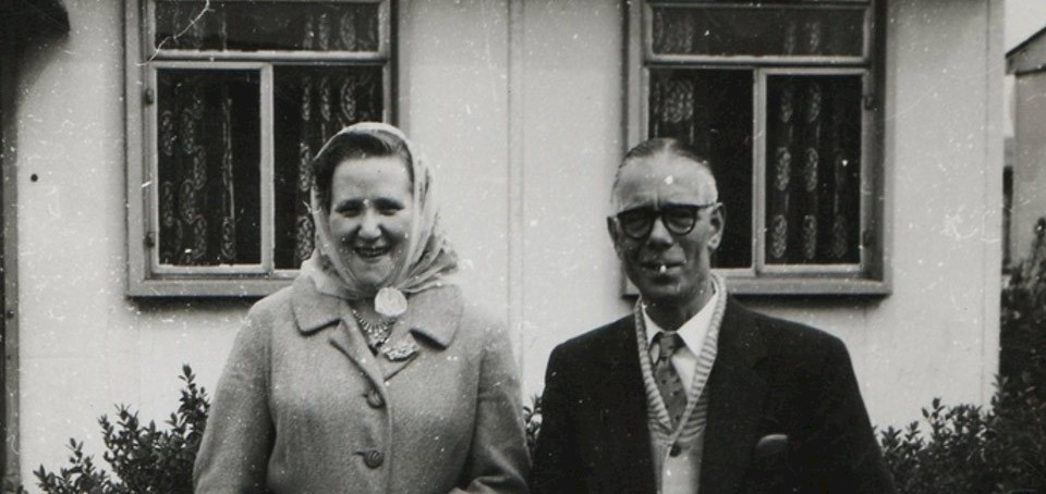 A couple standing outside a house, dressed smartly in mid‑20th‑century attire, pose for a photograph in front of their garden