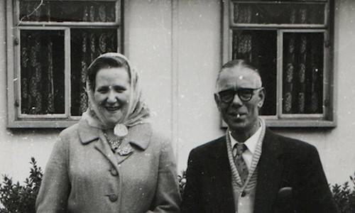 A couple standing outside a house, dressed smartly in mid‑20th‑century attire, pose for a photograph in front of their garden