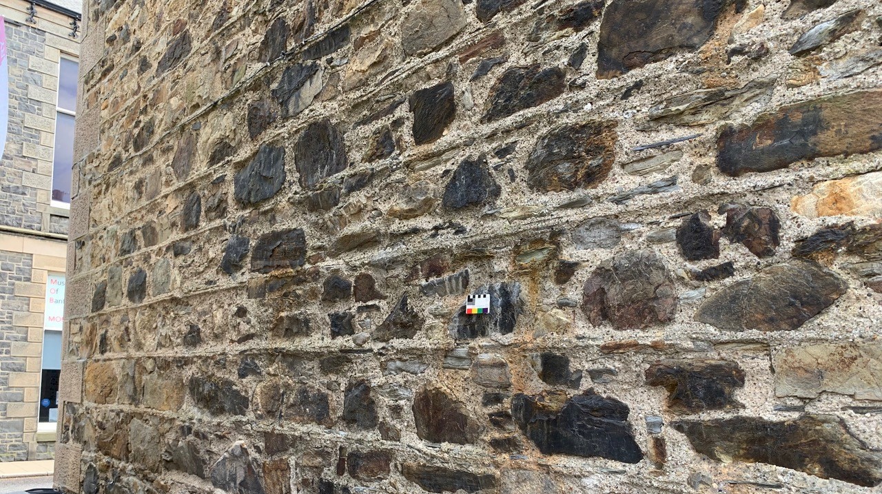 A traditional stone wall of a building. The building is pointed with lime and there's a small card placed on the wall to help identify the lime type.