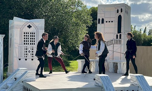 Five people on an outdoor stage pointing swords at each other