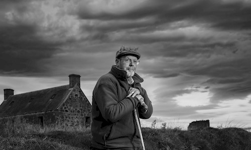 A man perching against a grass verge leaning on a stick with a bothy in the background