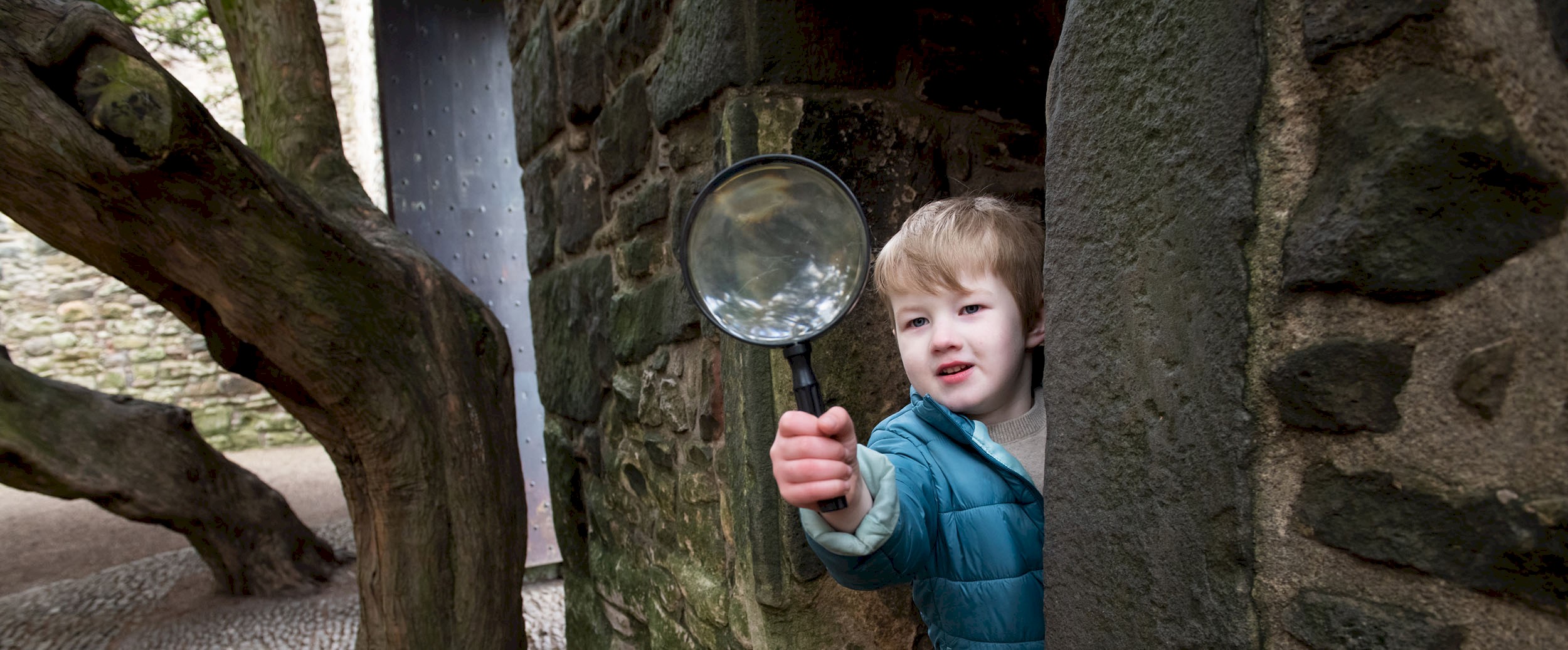 A boy peaking round from a large brick wall holding a magnifying glass
