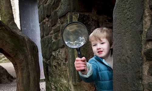 A boy peaking round from a large brick wall holding a magnifying glass
