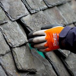 Close up of a gloved left hand of a restoration worker reaching out to touch an old fashioned slate roof. Some of the dark grey slates are not straight, with gaps in between.