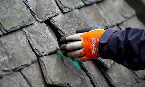 Close up of a gloved left hand of a restoration worker reaching out to touch an old fashioned slate roof. Some of the dark grey slates are not straight, with gaps in between.