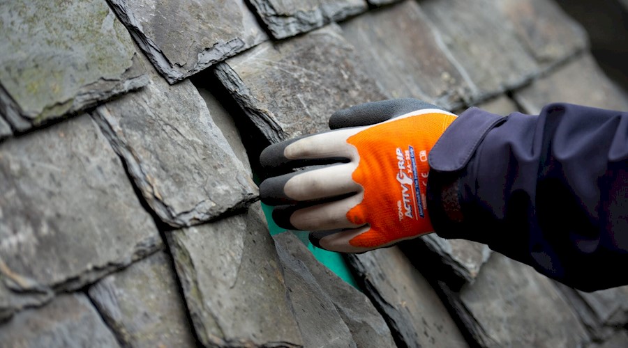 Close up of a gloved left hand of a restoration worker reaching out to touch an old fashioned slate roof. Some of the dark grey slates are not straight, with gaps in between.