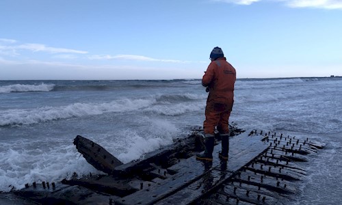 A person in overalls and wellingtons at the Sanday Shipwreck with big waves sweeping in