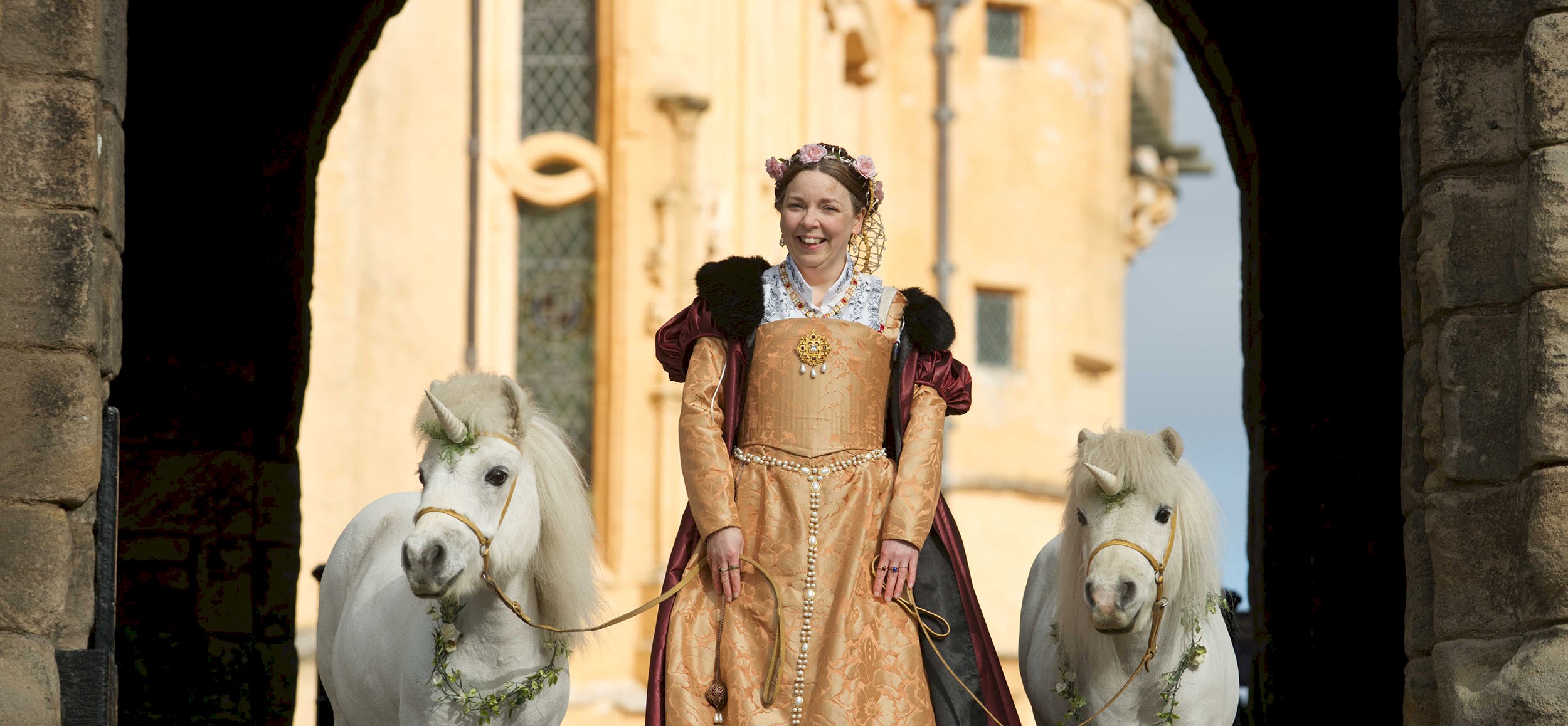 A woman dressed in period costume holding onto two ponies with unicorns on their heads
