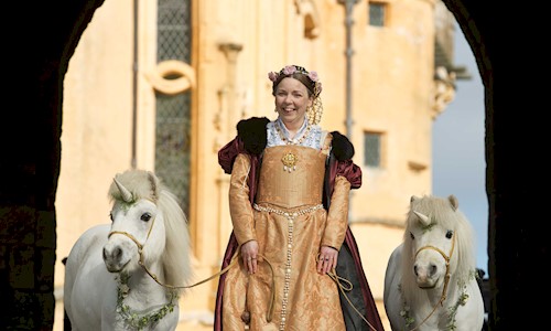 A woman dressed in period costume holding onto two ponies with unicorns on their heads
