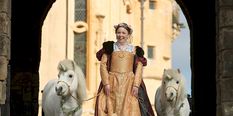 A woman dressed in period costume holding onto two ponies with unicorns on their heads