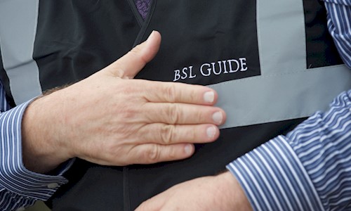 A close up of a mans hands. His right hand is flat on his chest and he is wearing a black tabard with the words BSL Guide printed on the chest.