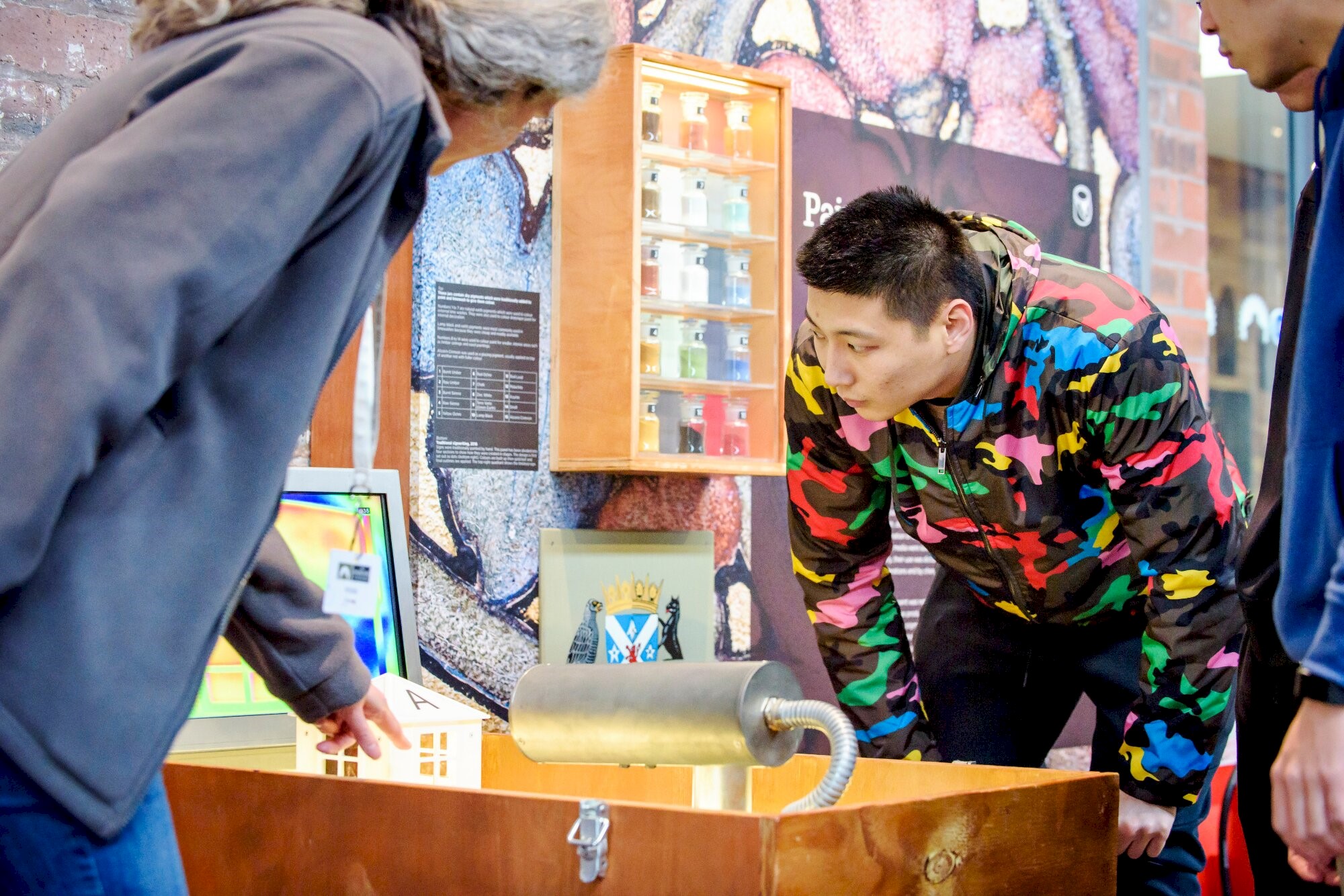 A group of people intently looking at a display in a museum setting; the display is a small white house indicating heat loss from a building. Another person is pointing something out as if explaining the display.