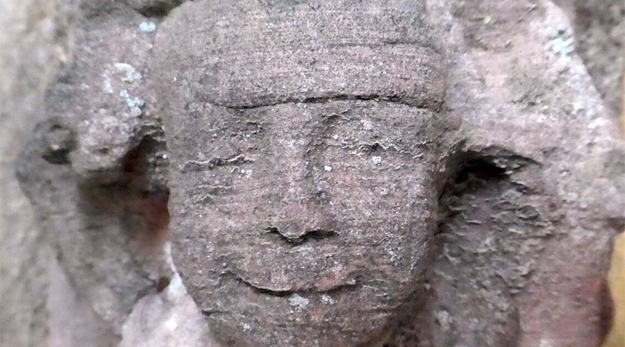 A close-up photo of a gargoyle type face on the side of a medieval abbey. The face is a human one, and is smiling, with the features eroded by the weather.