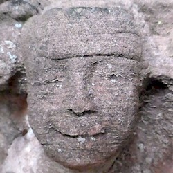 A close-up photo of a gargoyle type face on the side of a medieval abbey. The face is a human one, and is smiling, with the features eroded by the weather.