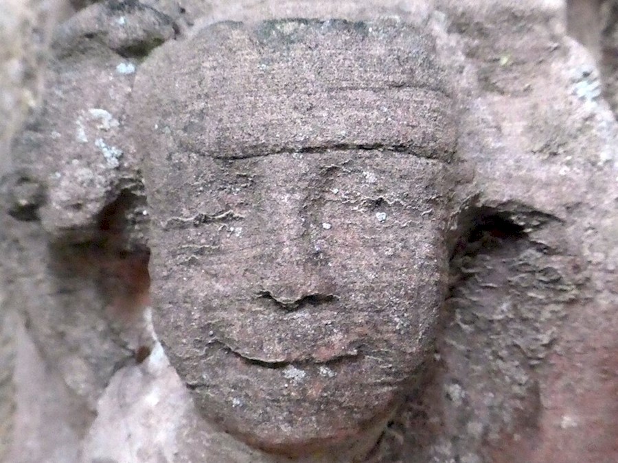 A close-up photo of a gargoyle type face on the side of a medieval abbey. The face is a human one, and is smiling, with the features eroded by the weather.