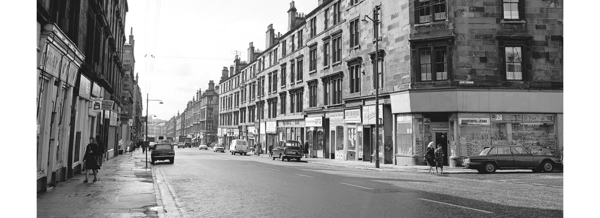 A black and white photo of a street in Scotland with shops at the bottom and tenement flats above, with cars parked and people walking along the pavements