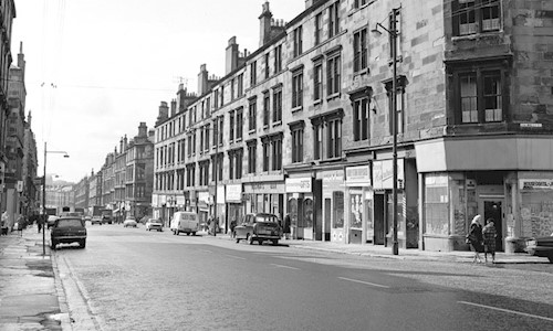 A black and white photo of a street in Scotland with shops at the bottom and tenement flats above, with cars parked and people walking along the pavements