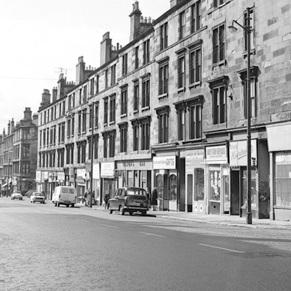 A black and white photo of a street in Scotland with shops at the bottom and tenement flats above, with cars parked and people walking along the pavements
