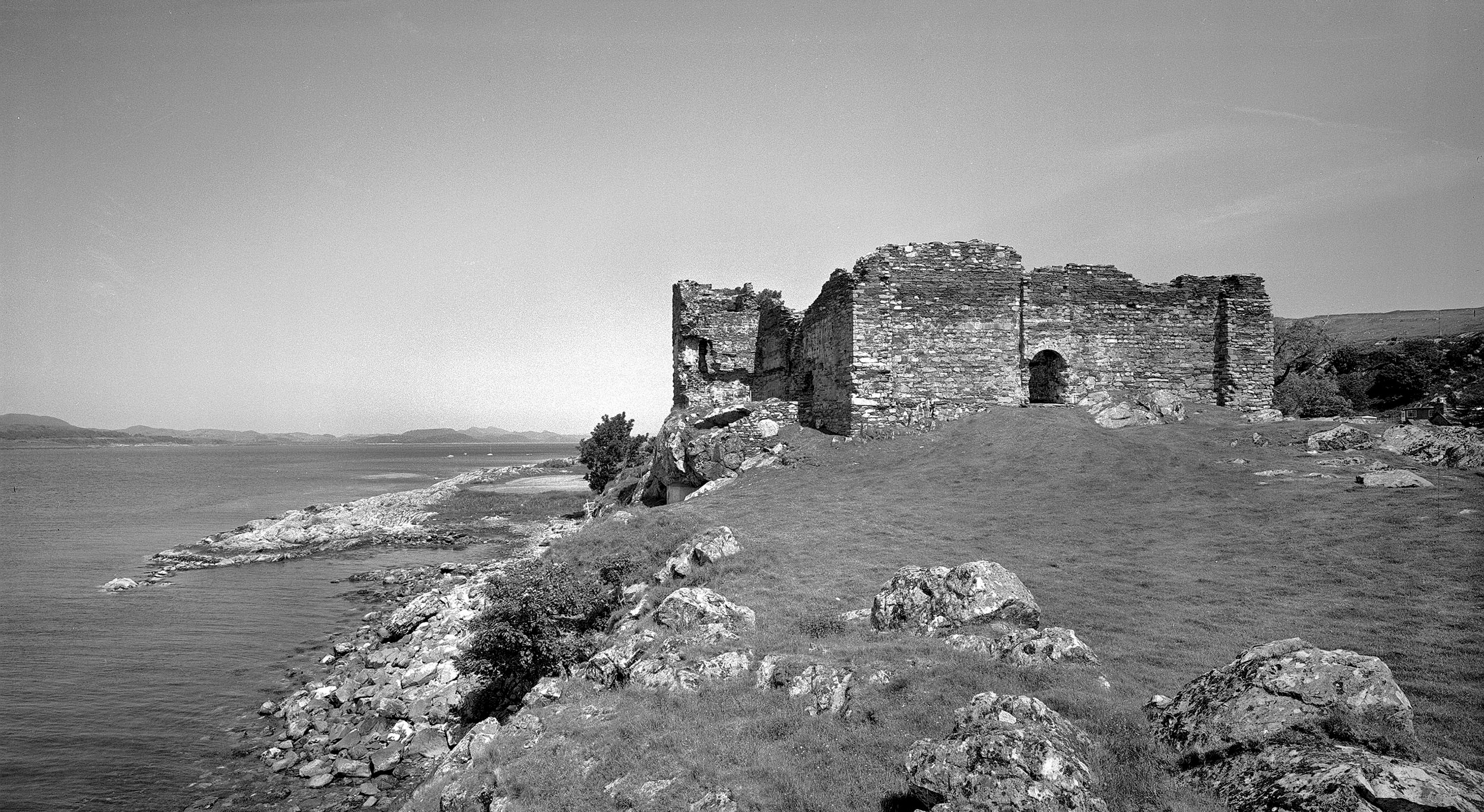 A black and white picture of castle ruins on top of a rocky hill with water below