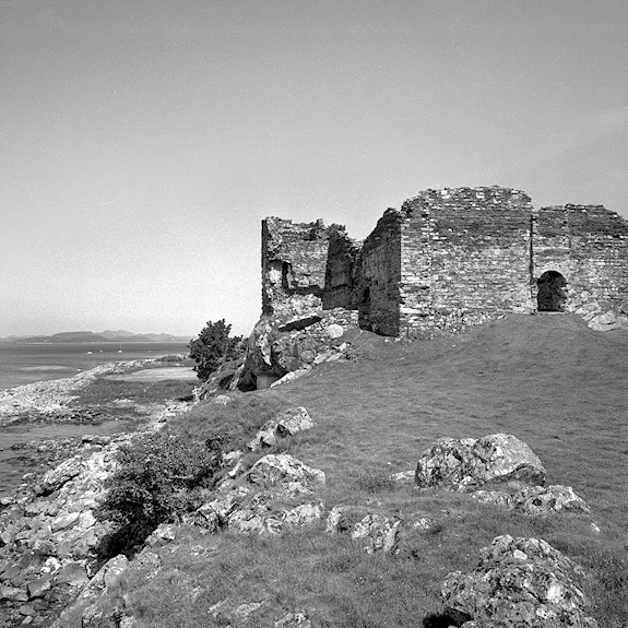 A black and white picture of castle ruins on top of a rocky hill with water below