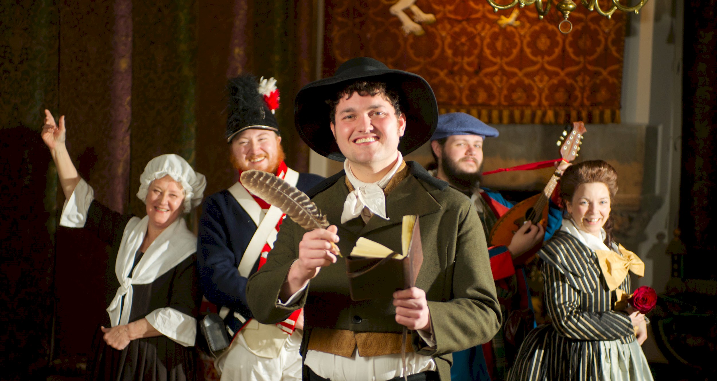 People in period costume, one as Rabbie Burns hodling a feather quill and a writing book. All of them have big smiles on their faces