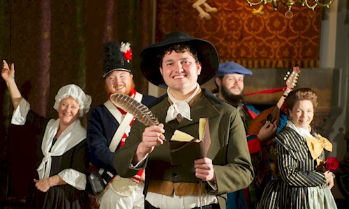 People in period costume, one as Rabbie Burns hodling a feather quill and a writing book. All of them have big smiles on their faces