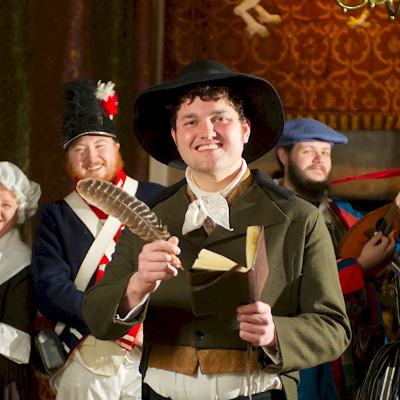 People in period costume, one as Rabbie Burns hodling a feather quill and a writing book. All of them have big smiles on their faces