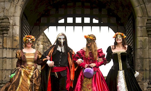 Four re-enactors in costume wearing masks with linked arms and walking under a portcullis gate 