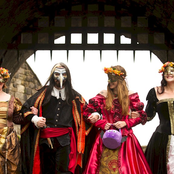 Four re-enactors in costume wearing masks with linked arms and walking under a portcullis gate 