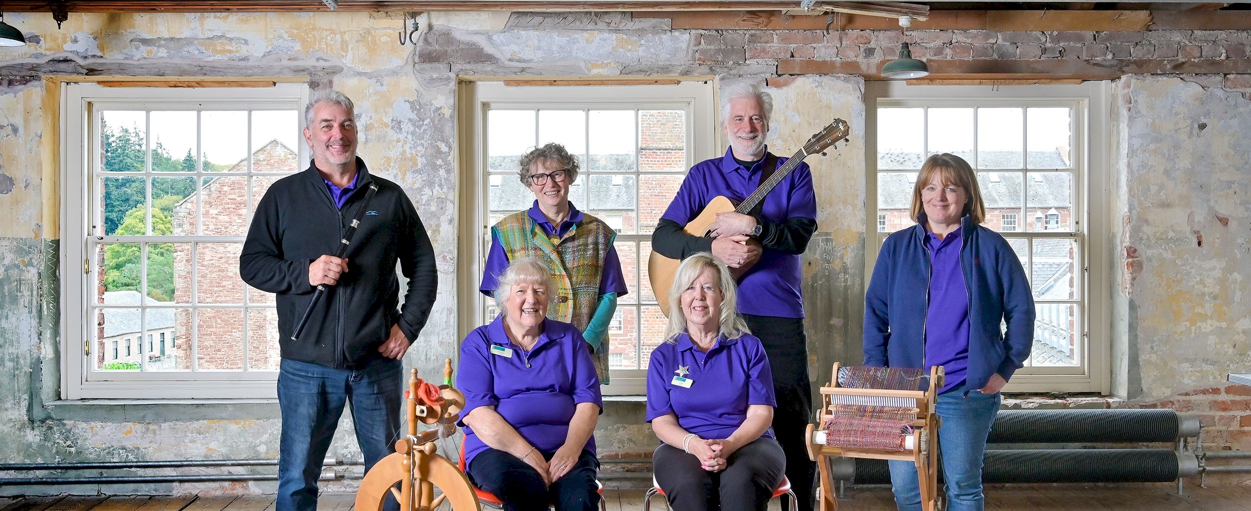 Six volunteers posing for a photograph all wearing branded purple polo shirts, one is holding a flute, another a guitar, and there's a loom and spinning wheel in front of them