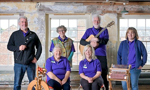 Six volunteers posing for a photograph all wearing branded purple polo shirts, one is holding a flute, another a guitar, and there's a loom and spinning wheel in front of them