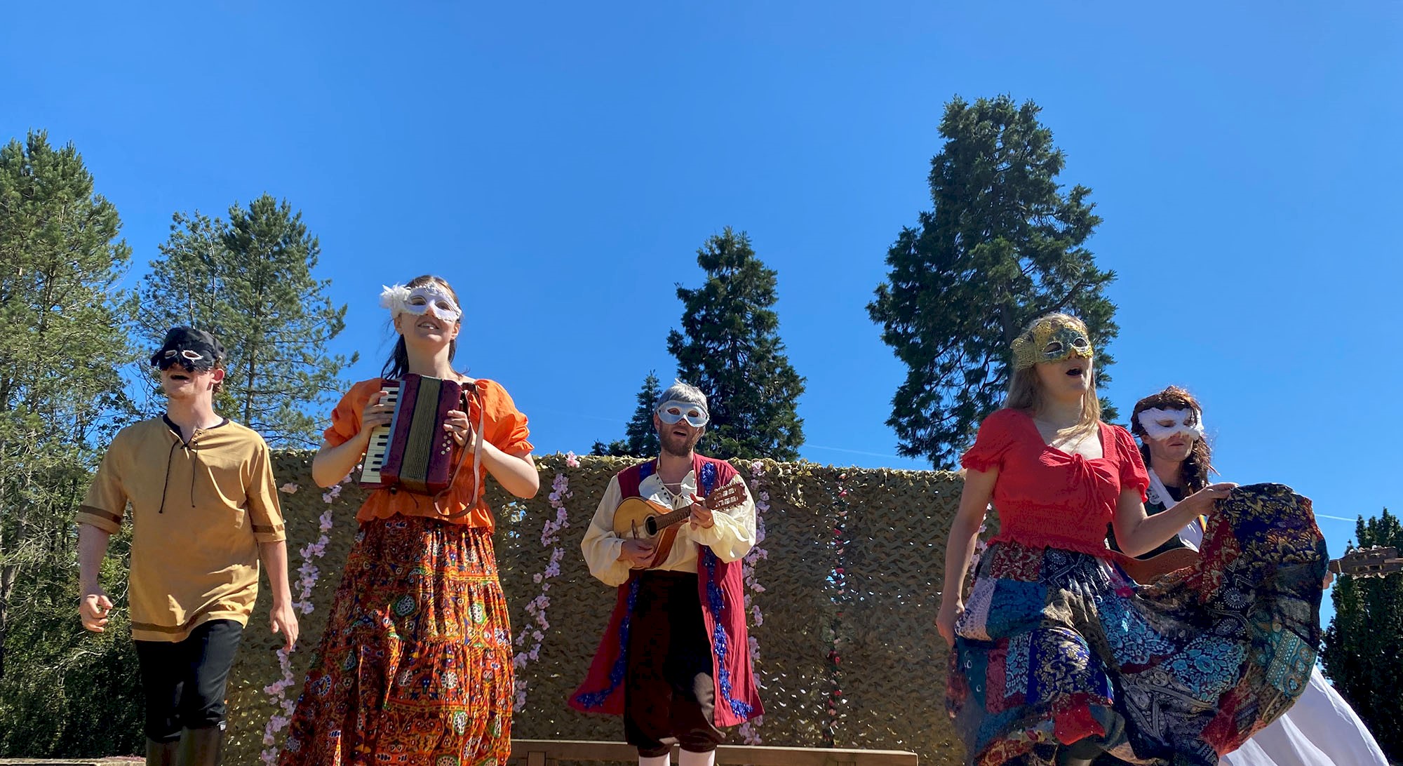 A group of costumed performers dressed in period clothing playing instruments and singing on an outdoor stage on a sunny day