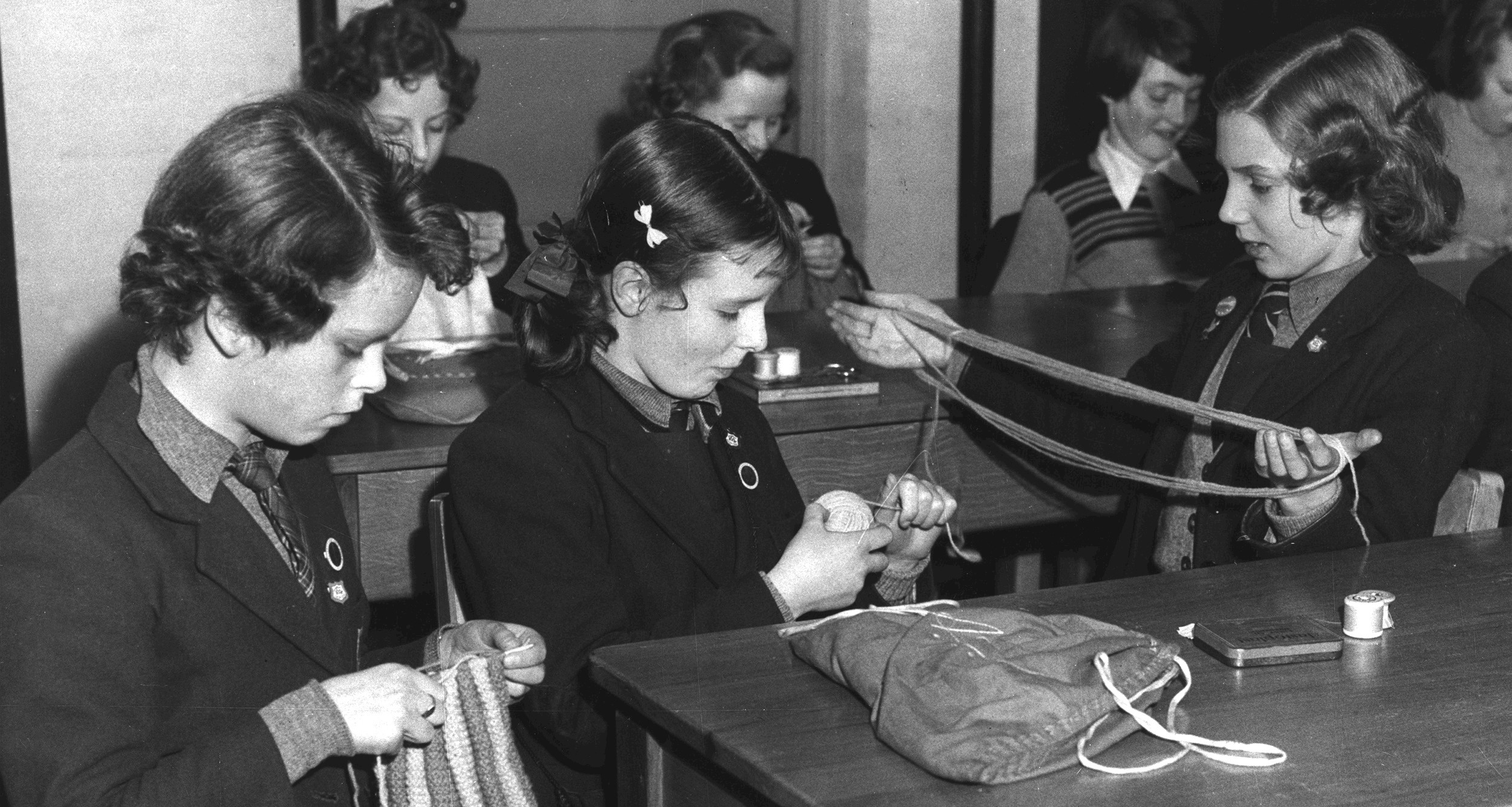 A black and white picture of girls sitting at wooden school desks some are knitting and some are holding the wool for the knitters