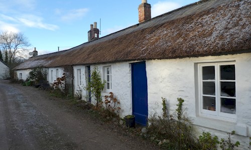 A row of whitewashed stone cottages with a long connected thatched roof. The doors of the cottages are blue, and they open directly onto a track road.