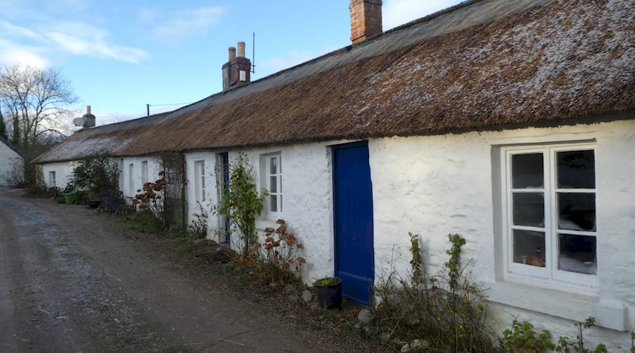 A row of whitewashed stone cottages with a long connected thatched roof. The doors of the cottages are blue, and they open directly onto a track road.