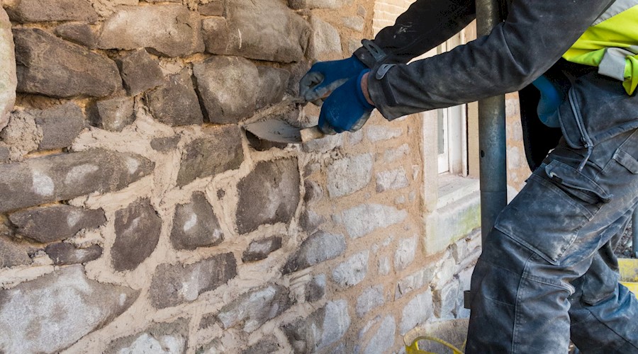 A person's arms working mortar in between cracks in an old stone wall of a building. They are wearing gloves and using a small, thin tool.