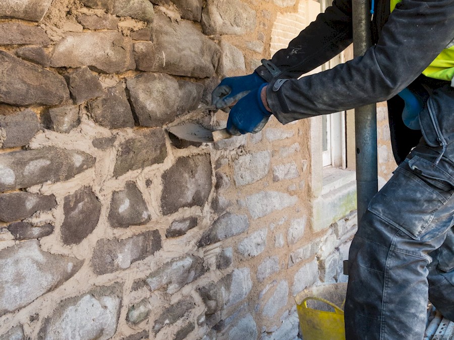 A person's arms working mortar in between cracks in an old stone wall of a building. They are wearing gloves and using a small, thin tool.