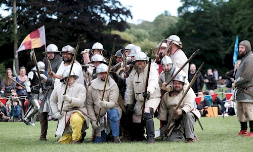 Multiple people in historic costume, wearing helmets and holding pole weapons at the ready to battle 