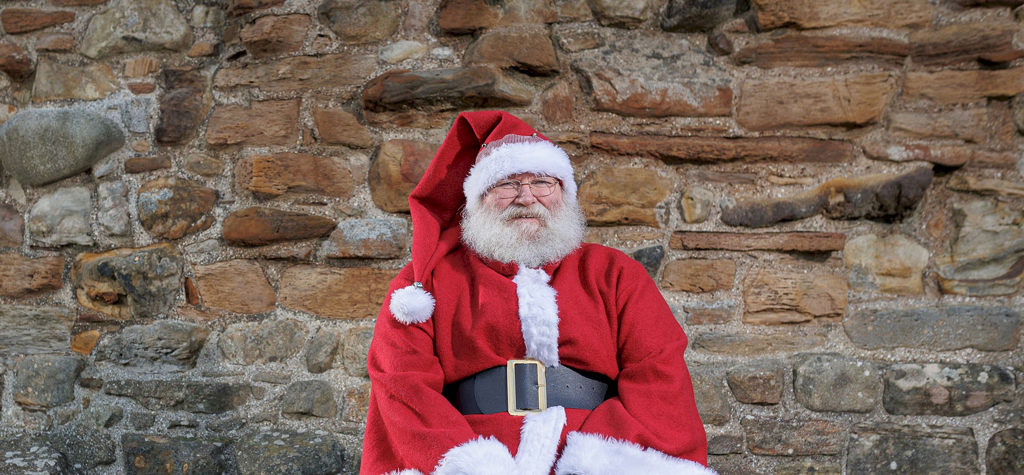 A man sitting on a bench in front of a high brick wall dressed as Santa Claus
